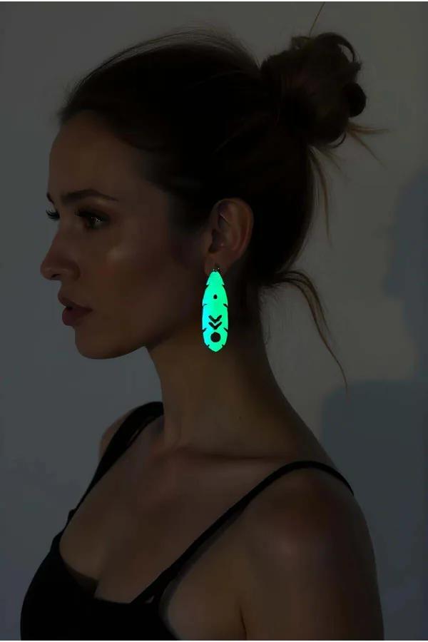 Glow-in-the-dark teardrop earrings emitting vivid cyan light with geometric chevron cutout pattern worn on ear