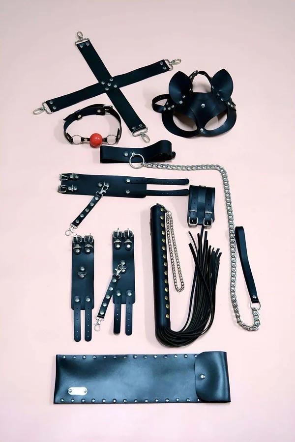 black faux leather restraint accessory kit flatlay showing hogtie bar wrist cuffs cat ear mask flogger chain leash and studde