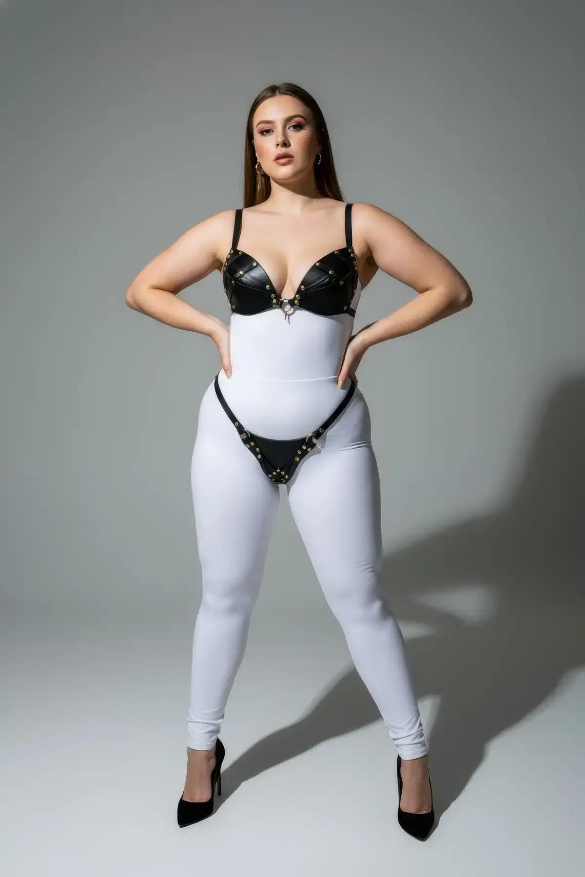 Black faux leather hip harness panel with gold studs and adjustable buckle closure worn over white leggings