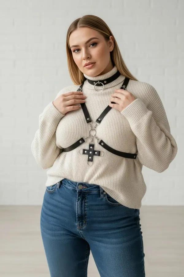 Black faux leather chest harness with silver O-ring choker and riveted cross pendant worn over cream knit sweater