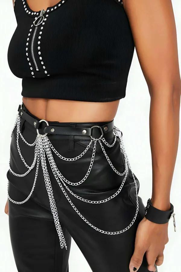 Black faux leather waist harness with cascading silver chain layers and O-ring studs worn over high-rise trousers