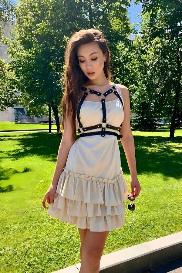 Black faux leather chest harness with gold O-rings worn over cream ruffled dress, adjustable strap cage design