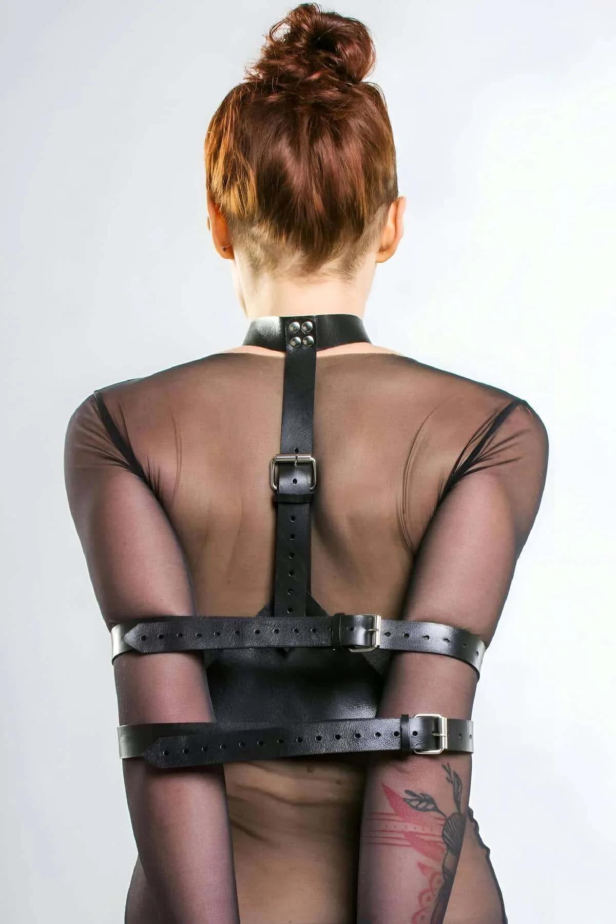 Black vegan leather back harness with wide arm cuffs and silver-tone buckles worn over sheer mesh top, back view