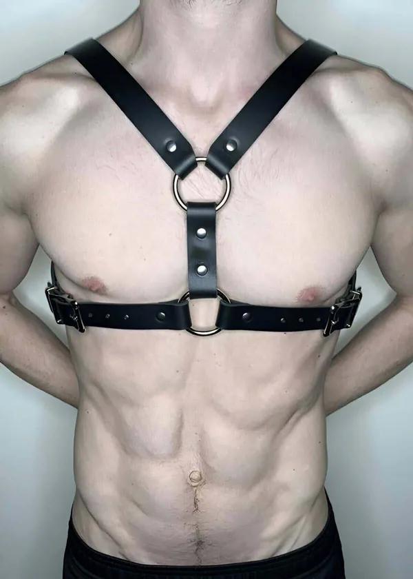 Black faux leather chest harness with two silver O-rings and adjustable side buckles worn on male torso