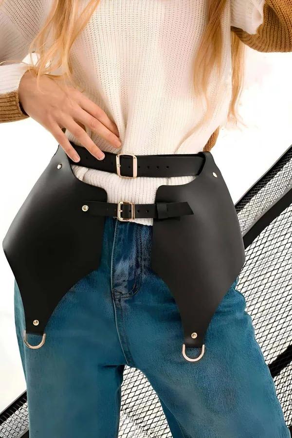 Black faux leather skirt harness belt with two gold buckle waist straps and D-ring panels worn over blue jeans