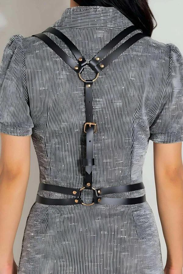 Black faux leather strappy body harness with gold-tone O-ring at upper back and adjustable waist band, worn over striped shir